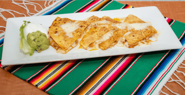 Golden quesadillas filled with melted cheese, served with creamy guacamole and sour cream on a rectangular plate over a vibrant Mexican tablecloth.