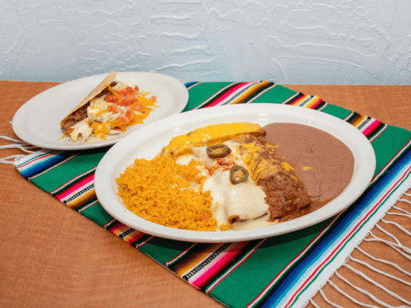 Traditional Mexican combination plate with cheesy enchiladas, refried beans, Mexican rice, and jalapeño slices, served with a crunchy beef taco on a festive green and red woven table runner.