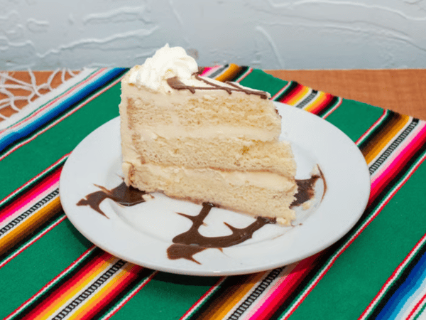 Slice of moist tres leches cake drizzled with chocolate syrup and topped with whipped cream, served on a white plate over a traditional Mexican tablecloth.