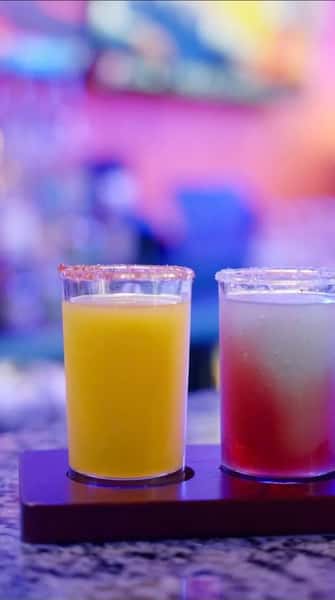 Colorful margarita flight featuring mango and watermelon flavors, served in salt-rimmed glasses on a wooden tray with a vibrant bar backdrop.