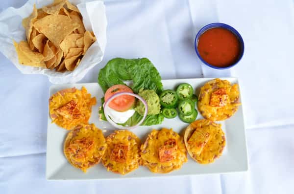 Appetizer platter of cheesy mini tostadas topped with grilled chicken, served with fresh jalapeños, guacamole, sour cream, and sliced vegetables, paired with tortilla chips and red salsa.