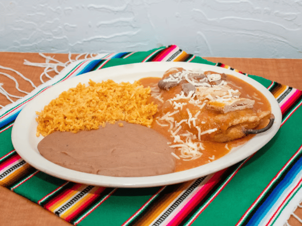 Chiles rellenos topped with red tomato sauce and shredded cheese, served with refried beans and Mexican rice on a festive woven table runner.
