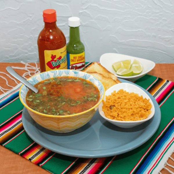 Colorful bowl of traditional Mexican caldo de res beef soup garnished with fresh cilantro, served with a side of fluffy Mexican rice, crispy tostadas, lime wedges, and hot sauces on a vibrant sarape tablecloth.