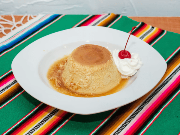 Classic Mexican flan with a caramel glaze, served with whipped cream and a cherry on a white plate over a vibrant sarape tablecloth.