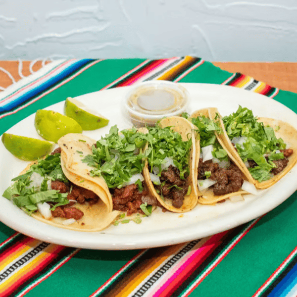 Authentic street-style Mexican tacos with grilled carne asada and al pastor, topped with fresh cilantro and onions, served with lime wedges and green salsa on a traditional sarape-patterned tablecloth.