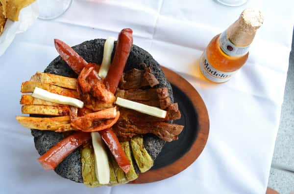 Sizzling molcajete filled with grilled shrimp, steak, sausage, nopal, and cheese strips, served in a traditional stone bowl with a cold bottle of Modelo beer.