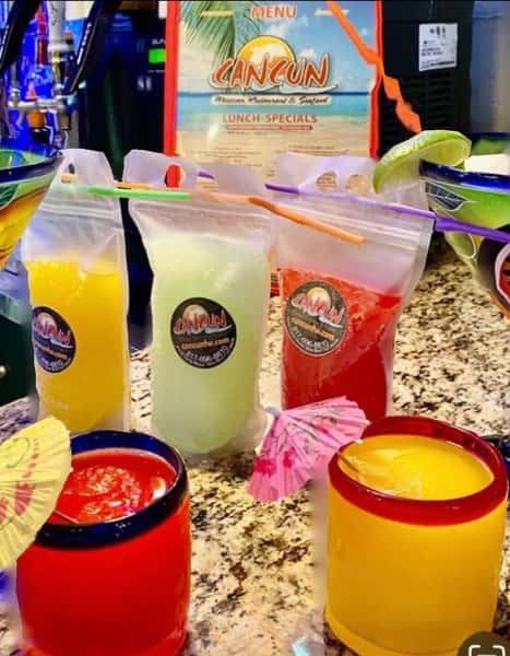 Colorful frozen margaritas in to-go pouches and rimmed glasses, featuring tropical flavors like mango, lime, and strawberry, served with cocktail umbrellas on a granite bar top at Cancun Mexican restaurant.