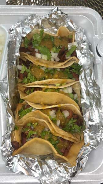 Takeout order of carne asada tacos served in a foil-lined container, topped with green salsa, chopped onions, and cilantro, showcasing authentic to-go Mexican street tacos.