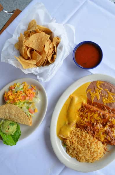 Mexican combination plate with cheesy enchiladas, refried beans, and seasoned rice, served with a crispy beef taco topped with lettuce and shredded cheese, guacamole, and a basket of tortilla chips with salsa.