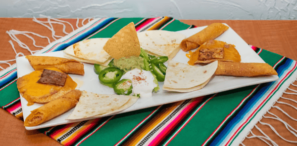 Mexican appetizer platter with mini quesadillas, taquitos, and tostadas topped with melted cheese and beef, served with sliced jalapeños, sour cream, guacamole, and a crispy tortilla chip centerpiece.