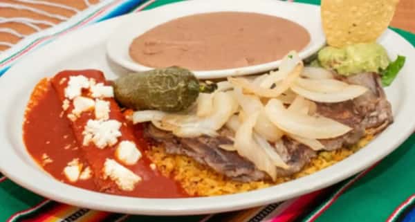 Traditional Mexican plate featuring grilled beef steak with sautéed onions, red enchiladas topped with queso fresco, refried beans, Mexican rice, and guacamole with a tortilla chip.