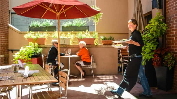 Outdoor patio dining with two women seated at a table and a server carrying two plates.