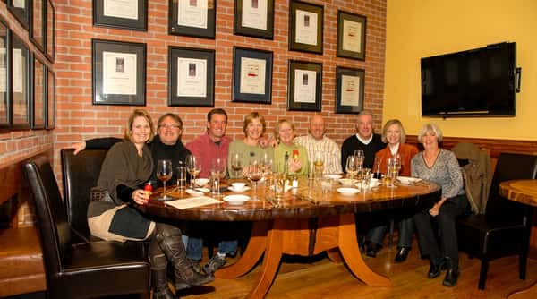 Nine adults seated around a large wooden table with wine glasses and plates, against a brick wall decorated with framed certificates.