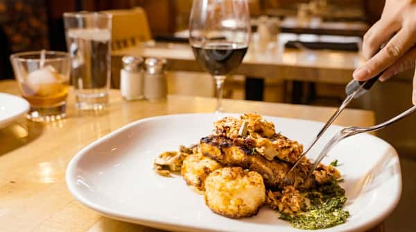 Grilled fish with crunchy topping, two potato cakes, and green pesto. A hand cuts into the fish.