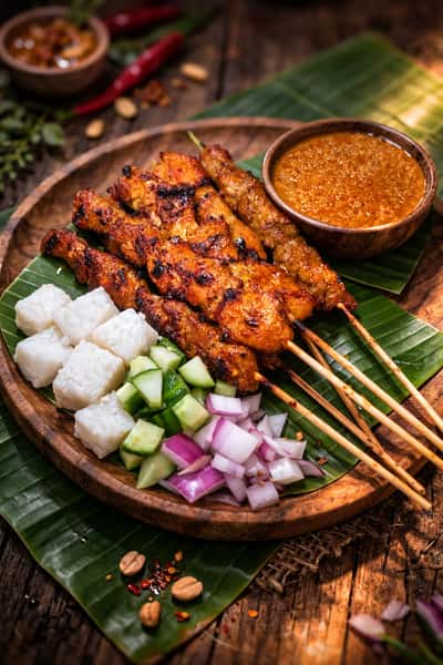 Grilled Malaysian beef satay skewers served with peanut sauce, rice cakes, fresh onions, and cucumber on a banana leaf platter.