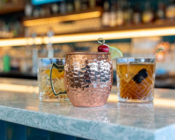 Two cocktails in patterned glasses on a bar, one in a copper mug style cup.