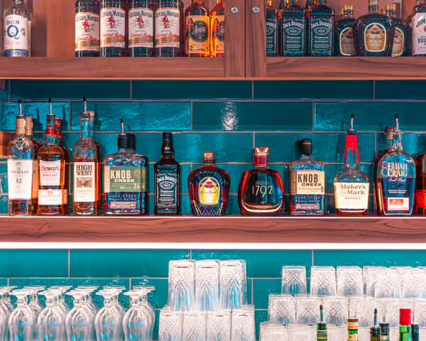 Row of liquor bottles lined up on a shelf against a teal wall.