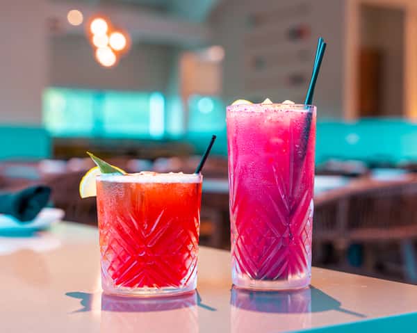 Two colorful cocktails in tall glasses on a table in a restaurant.