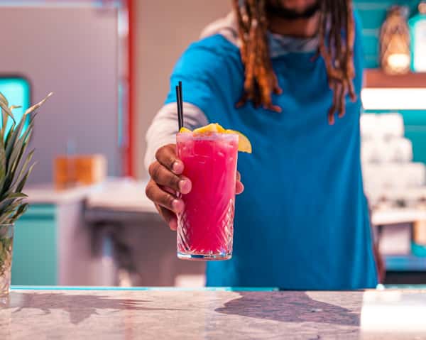 Hand holding a bright pink cocktail with a straw in a bar setting.