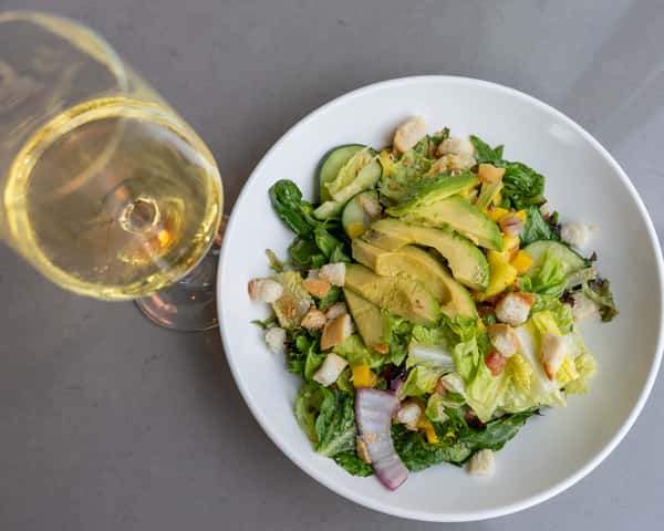 Salad topped with sliced avocado on a white plate, with a wine glass nearby.