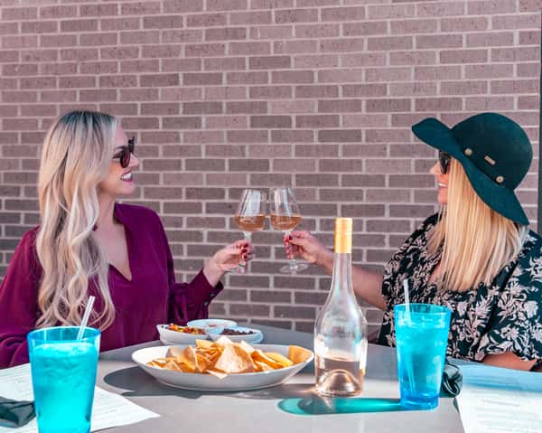 Two women seated outdoors clinking wine glasses in front of a brick wall.