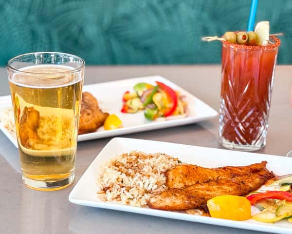 Glass of beer on a table next to plated food and a red drink in the background.