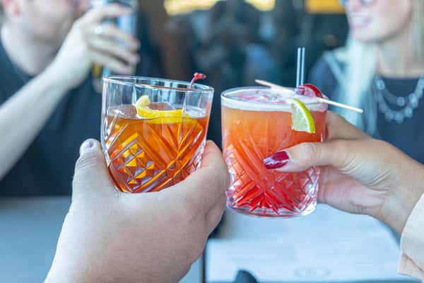 Two hands holding cocktails clinking glasses in a restaurant setting.
