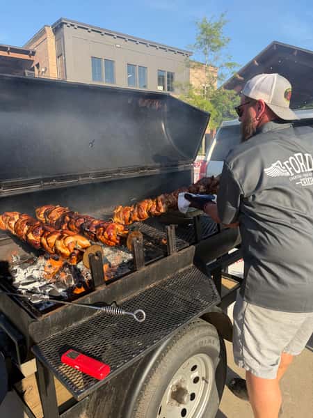 Person tending a rotisserie grill with whole chickens cooking over charcoal.