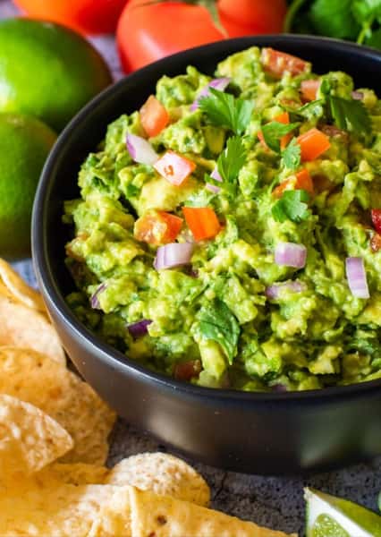 Freshly made chunky guacamole in a black bowl, garnished with diced tomatoes, red onions, and cilantro—served with tortilla chips and surrounded by limes and tomatoes, ideal for authentic Mexican appetizers.