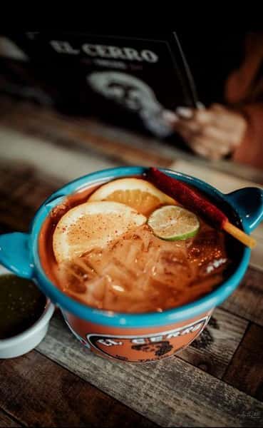 Vibrant Mexican cocktail served in a traditional clay mug, garnished with lime, orange slices, and a tamarind candy stick—perfect for a refreshing, citrus-forward drink at a Mexican cantina or taqueria.