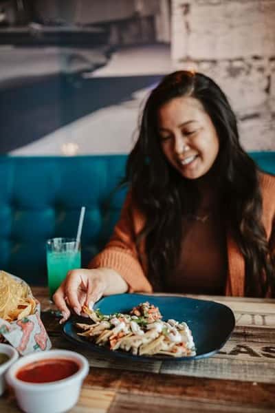 Smiling woman enjoying loaded nachos with salsa and a vibrant aqua-blue drink in a cozy Mexican restaurant booth—ideal for showcasing lively, casual dining experiences.