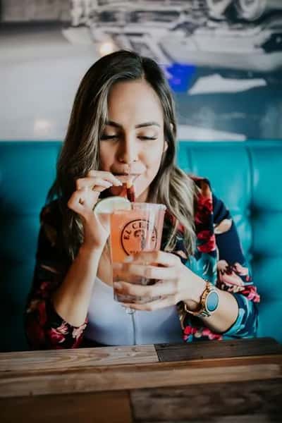 Woman sipping a refreshing pink Mexican cocktail garnished with lime and tamarind candy, seated in a vibrant restaurant setting—ideal for promoting festive drinks and cantina vibes.