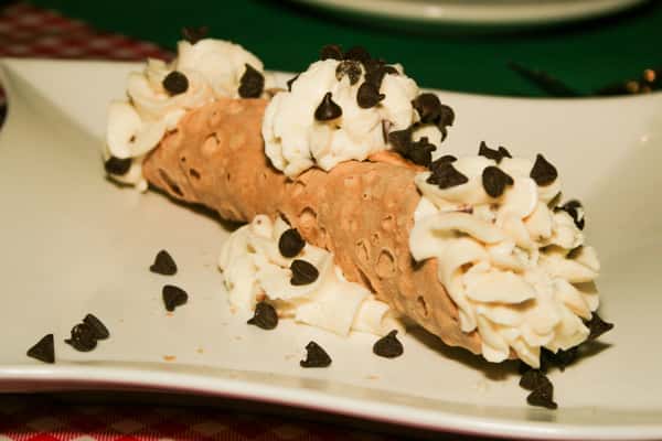 A chocolate cannoli filled with rich cream. The pastry is topped with several dollops of whipped cream and sprinkled generously with mini chocolate chips, served on a white plate.