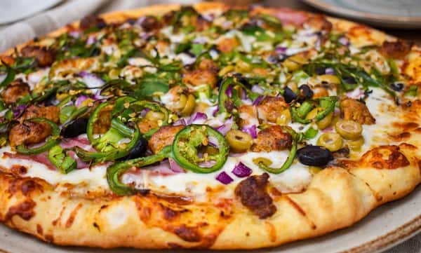 Close-up of a freshly baked specialty pizza topped with grilled chicken, jalapeños, red onions, green and black olives, melted mozzarella, and a golden hand-tossed crust.