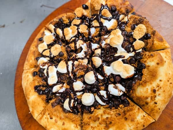 Dessert pizza topped with toasted marshmallows, chocolate chips, cookie crumbles, and chocolate drizzle on a freshly baked sweet crust.