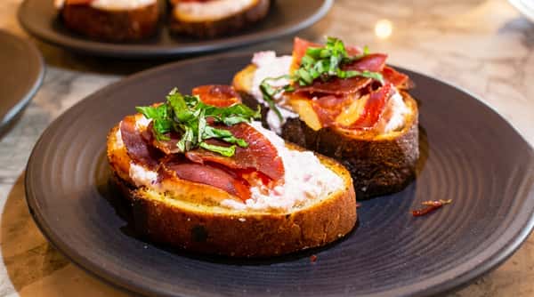 Toasted bread topped with creamy cheese, thin slices of meat, and fresh basil leaves on a black plate.