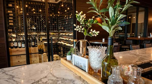 Wine cellar behind a glass wall, with a marble bar top and decorative bottles and plants in the foreground.