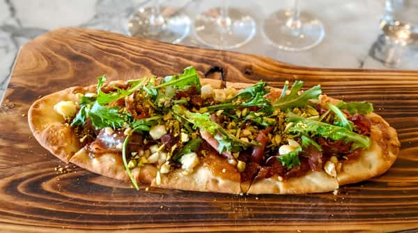 Flatbread with prosciutto, arugula, pistachios, cheese, and balsamic glaze on a wooden board.