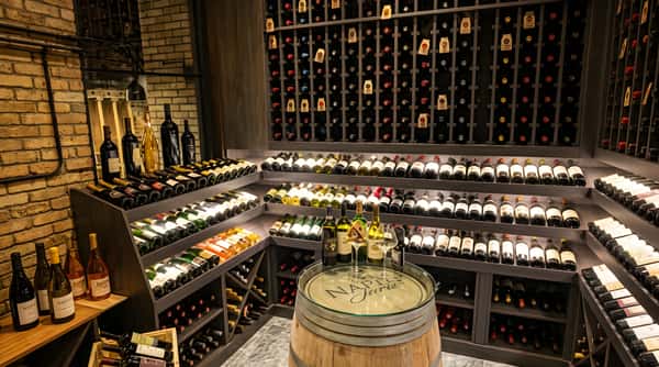 A large wine cellar with bottles displayed on wooden racks and shelves, featuring a barrel table.