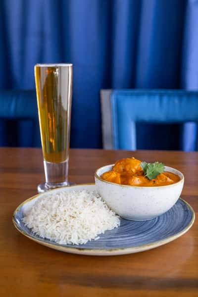 A bowl of Indian curry with rice and a tall glass of beer in the background.