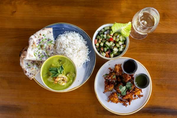 A platter featuring a bowl of green curry with vegetables, a serving of white rice, naan bread, a side salad with diced vegetables, and a plate of fried snacks accompanied by dipping sauces.