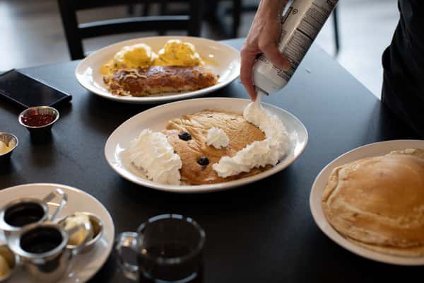 Kid-friendly pancake decorated with whipped cream and blueberries to resemble a smiling face, being topped with fresh whipped cream at the table — playful and inviting breakfast at a family-oriented diner.