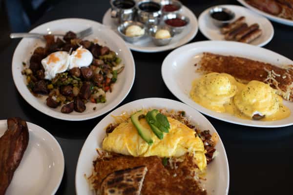 Hearty diner breakfast featuring a fresh avocado omelette with hash browns and grilled toast, classic eggs Benedict with hollandaise sauce, and a roasted vegetable breakfast hash with poached eggs — served with assorted condiments for a full morning feast.