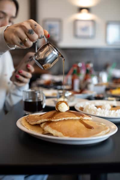 Stack of fluffy pancakes with melting butter being drizzled with warm maple syrup, served at a cozy American diner — a comforting breakfast favorite enjoyed in a casual setting.
