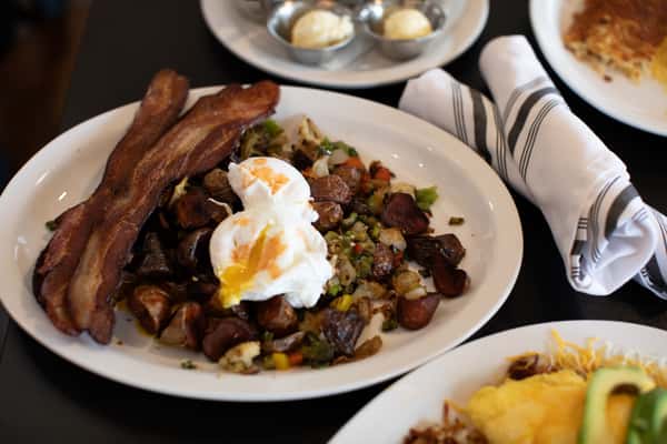 Diner-style breakfast plate featuring crispy bacon, rustic potato hash with peppers and onions, and two perfectly poached eggs with runny yolks — a bold and savory morning meal.