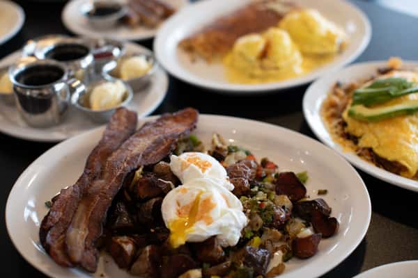 Crispy bacon strips served with a rustic breakfast hash of diced potatoes, peppers, and onions, topped with two perfectly poached eggs. A rich and satisfying American breakfast platter at a cozy diner.