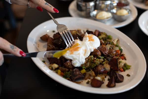 Hands with red nail polish cut into poached eggs on a plate of breakfast potatoes mixed with vegetables, the egg yolk running over the dish on a white plate.