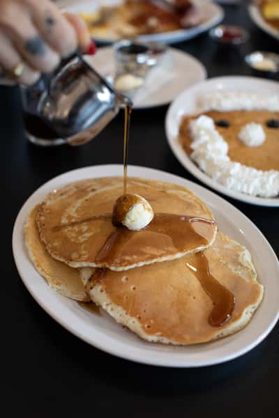 Stack of fluffy pancakes topped with butter and generously drizzled with maple syrup, with a fun whipped cream face pancake in the background — capturing a mix of playful and traditional breakfast offerings at a welcoming diner.