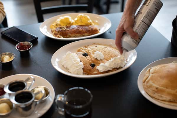 Smiley face pancake decorated with whipped cream and blueberries, being topped with extra whipped cream at the table — a playful, kid-friendly breakfast option at a family diner.