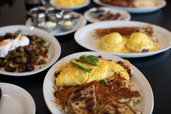 Southwestern-style omelette loaded with diced vegetables and cheese, topped with avocado slices and served with hash browns and grilled toast. Background plates include eggs Benedict, breakfast sausage, and a roasted potato hash for a filling diner breakfast.